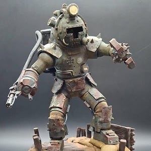 Könnte beinhalten: Eine verwitterte, olivgrüne gepanzerte Figur mit einem Tiefseetaucherhelm mit T-förmigem Visier, einem Eisernen Kreuz auf der Brust, stacheligen Schulterklappen und einer mechanischen Greifhand, stehend auf einem Sockel mit Sandsäcken und Holzplanken.