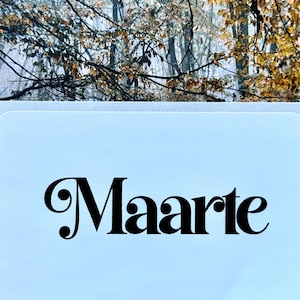 May include: A rectangular white sign featuring the word Maarte in a bold black serif font. The sign is positioned against a backdrop of autumn trees with yellow and brown leaves, creating a contrast between the clear text and the foliage.