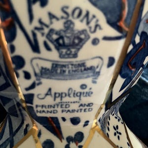 Op de afbeelding: Een keramisch object met een blauw-wit bloemmotief en gouden rand. Het fabrieksmerk toont Mason's, Ironstone, Made in England, Appliqué, Printed and Hand Painted onder een kroonicoon op een crèmekleurige achtergrond.
