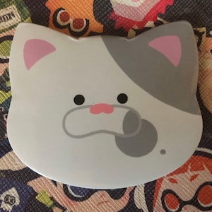 Può includere: Un viso di gatto bianco e grigio con orecchie e naso rosa, stampato su uno sfondo bianco. Il gatto ha occhi neri e una macchia grigia sulla fronte.