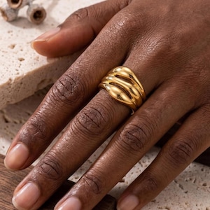May include: A polished gold ring featuring a multi-layered, rounded band design with three distinct, smooth, and curved segments that wrap around the finger, creating a continuous, fluid, and reflective metallic surface across the entire piece.