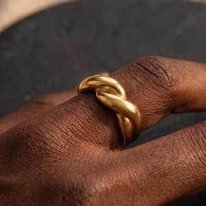 May include: A polished gold ring featuring a thick, twisted rope design. The band consists of two intertwined metal strands that create a continuous, rounded knot pattern. The surface of the gold metal is smooth and reflective, catching the light.