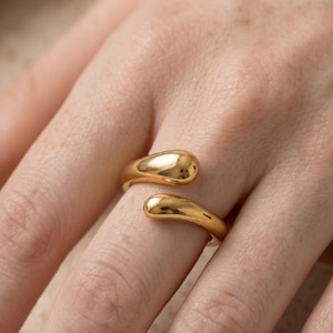 May include: A gold-toned open ring featuring two smooth, rounded, teardrop-shaped ends that curve towards each other. The metal surface is highly polished and reflective, creating a sleek, continuous band design that wraps around the finger.