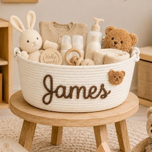 May include: A white woven rope basket featuring the name James in brown script lettering and a small brown teddy bear face appliqué. The basket is filled with baby items including a plush rabbit, a teddy bear, bottles, and a wooden ring.