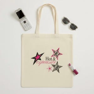 Puede incluir: Una bolsa de tela color crema con el texto negro Hot & Expensive. El diseño incluye varias estrellas en rosa intenso sólido y patrones de cebra en blanco y negro. La bolsa está acompañada por un teléfono plegable plateado, gafas de sol y un labial.
