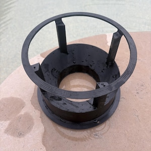 May include: A black circular plastic stand featuring a wide base, a central opening, and a top ring supported by four vertical struts. The surface of the object shows small water droplets, indicating a wet or recently submerged condition.