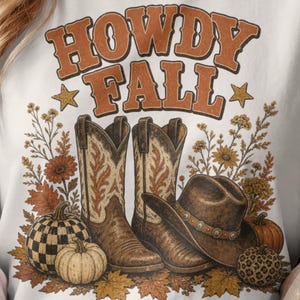 May include: A white t-shirt featuring a graphic print with the text HOWDY FALL in orange letters. The design includes two cowboy boots, a brown cowboy hat, pumpkins, and autumn leaves. The pumpkins have checkered and leopard print patterns.