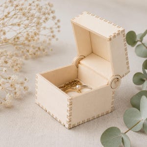 Puede incluir: Una pequeña caja de madera de color claro con tapa abatible y esquinas con juntas de dedos. La caja está abierta, revelando un collar de cadena de tono dorado y un anillo de tono plateado con una perla blanca dentro del recipiente cuadrado.
