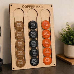 Pode incluir: Um suporte para cápsulas de café em madeira clara com o texto COFFEE BAR gravado no topo. Possui três ranhuras verticais que contêm quinze cápsulas de café Nescafé Dolce Gusto nas cores castanho, preto e laranja dispostas em filas.