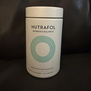 May include: A white cylindrical bottle of Nutrafol Women's Balance hair growth nutraceutical dietary supplement. The label features black text and a teal circular graphic. The bottle contains 120 capsules and has a matching white lid.