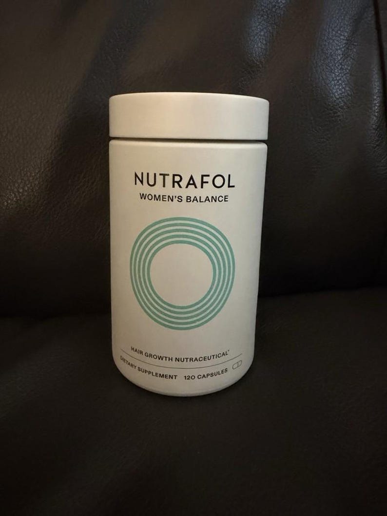 May include: A white cylindrical bottle of Nutrafol Women's Balance hair growth nutraceutical dietary supplement. The label features black text and a teal circular graphic. The bottle contains 120 capsules and has a matching white lid.