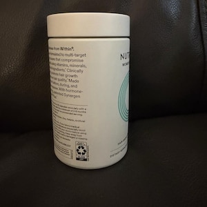 May include: A white cylindrical supplement bottle with a flat lid. The label features black text detailing product information, including the phrase Wellness from Within, and a teal circular graphic. The bottle is made of glass and includes a recycling icon.