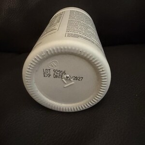 May include: A white cylindrical plastic bottle with a ribbed base. The bottom of the bottle features printed black text that reads LOT 92916 and EXP DATE 09/2027, along with a small recycling symbol containing the number 2.