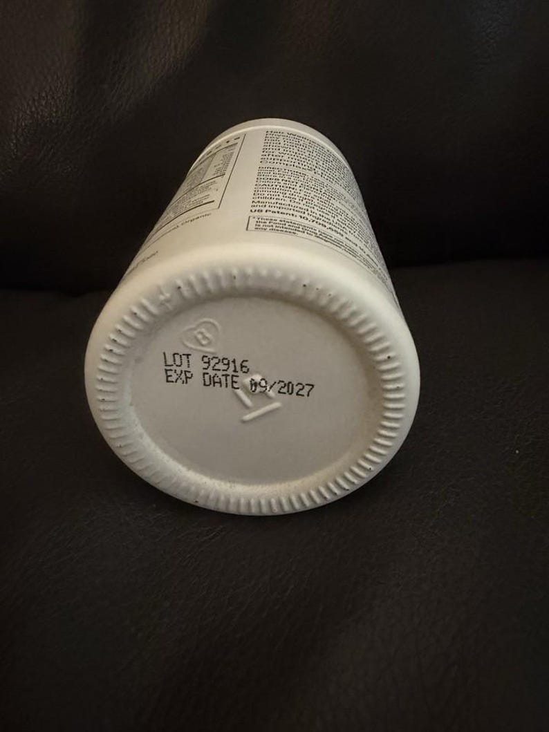 May include: A white cylindrical plastic bottle with a ribbed base. The bottom of the bottle features printed black text that reads LOT 92916 and EXP DATE 09/2027, along with a small recycling symbol containing the number 2.