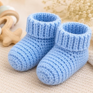 Könnte beinhalten: Ein Paar hellblaue Baby-Schühchen mit gehäkelter Textur. Die Schühchen verfügen über ein dickes, geripptes Umschlagbündchen am Knöchel und ein abgerundetes Zehendesign, das ein durchgehendes und gleichmäßiges Maschenbild aufweist.