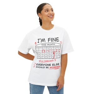 Può includere: Una t-shirt bianca con il testo nero I'M FINE sopra un grafico di un calendario con cerchi rossi attorno alle date dall'8 al 12. Sotto il calendario, il testo rosso recita ON THESE DAYS e il testo nero EVERYONE ELSE SHOULD BE WORRIED.