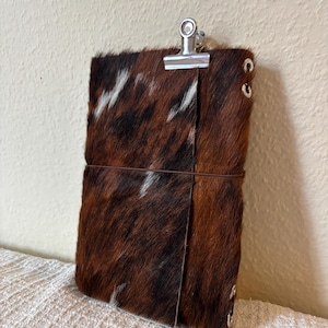 Può includere: Una copertina per taccuino rettangolare caratterizzata da una texture in pelle bovina nei colori marrone, nero e bianco. La copertina è fissata con un sottile elastico marrone e presenta occhielli in metallo argentato sul lato, con una clip.