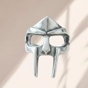 Op de afbeelding: Een zilverkleurige metalen ring in de vorm van een gladiatorenmasker. De ring heeft een puntig voorhoofdgedeelte, twee rechthoekige ooguitsparingen, een centrale neusbrug en twee verticale staven die naar beneden lopen vanaf de mond met een verweerde afwerking.