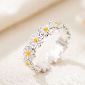 May include: A silver-toned metal ring featuring a continuous band of daisy flower motifs. Each flower has white petals and a bright yellow center. The ring has an open-ended design, allowing for adjustment, and displays a polished metallic finish.
