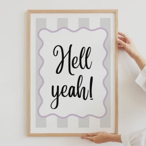 May include: A rectangular wall art print in a light wood frame featuring a white background with vertical grey stripes. A central lavender wavy-edged rectangle contains the black cursive text Hell yeah! centred in the middle of the frame.