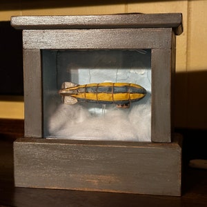 Puede incluir: Una vitrina de madera con frente de vidrio, que muestra un modelo amarillo y negro de una aeronave flotando sobre un fondo de nubes blancas.