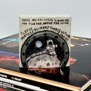 May include: A small, square, hand-painted artwork depicting an astronaut on the moon, viewed through a circular window. The artwork includes the text "HERE AM I FLOATING 'ROUND MY TIN CAN FAR ABOVE THE MOON PLANET EARTH IS BLUZ AND THERE'S NOTHING CAN DO."
