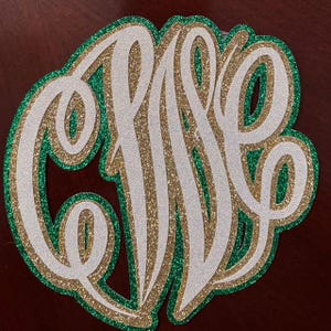 Può includere: Un monogramma a strati composto da tre forme circolari sovrapposte. Lo strato inferiore è glitter verde, quello centrale è glitter oro e quello superiore è glitter bianco. Il design mostra lettere corsive stilizzate e interconnesse.