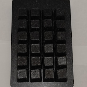 May include: A rectangular black macro pad featuring a grid of sixteen square keys arranged in four rows and four columns. The device has a matte finish with a visible textured surface on each keycap and the surrounding frame, providing a uniform look.