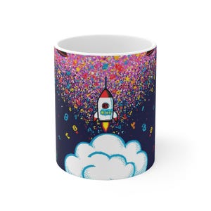 Könnte beinhalten: Ein weißer Keramikbecher mit dunkelblauem Hintergrund, auf dem eine Rakete von einer weißen Wolke abhebt. Die Rakete ist von einer lebendigen Explosion aus bunten Zahlen und Buchstaben umgeben, was ein dynamisches und detailreiches Muster erzeugt.