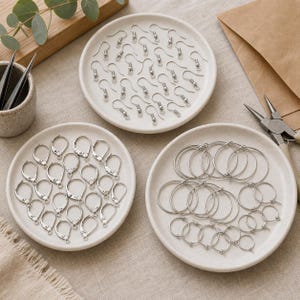 May include: A collection of silver-toned jewelry findings arranged on three small, round, off-white ceramic plates. The set includes fishhook earring wires, lever-back earring components, and circular hoop earring findings, accompanied by metal pliers.
