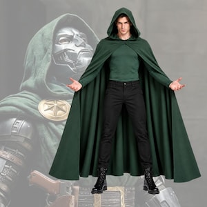 May include: A dark green hooded cloak worn over a matching green long-sleeve shirt and black trousers. The cloak features a circular gold-toned brooch at the shoulder. A large, faded graphic of a metallic masked figure is visible in the background.