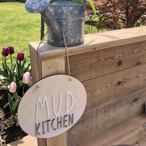 Op de afbeelding: Een gieter van gegalvaniseerd metaal gevuld met witte en lichtroze tulpen, met een hangend ovaal houten bordje met de tekst MUD KITCHEN in witte en zwarte letters, opgehangen aan een dun koord aan het handvat van de gieter.