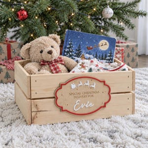 May include: A light-coloured wooden crate featuring a decorative plaque with the text "SPECIAL CHRISTMAS DELIVERY FOR Evie" and a silhouette of Santa and reindeer. Inside the crate sits a tan teddy bear with a red plaid bow and a storybook.