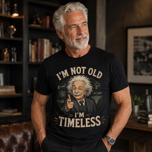 May include: A black t-shirt featuring a graphic of Albert Einstein pointing forward with the text "I'M NOT OLD I'M TIMELESS" in a vintage font. The design includes scientific formulas like E=mc2 and a lightbulb icon on a dark circular background.