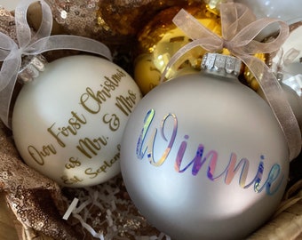 May include: Two Christmas ornaments, one white with gold lettering that reads "Our First Christmas as Mr. & Mrs. Est. September" and one silver with iridescent lettering that reads "Winnie".