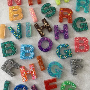 May include: A collection of colorful resin letters and shapes, each embedded with glitter. The letters are in various colors, including red, green, blue, orange, and purple. Some letters have gold leaf accents.