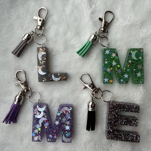 May include: Four resin letter keychains spelling "LMEE". The letters are filled with glitter and celestial designs, including moons and stars. Each keychain has a different color tassel and a silver clasp.