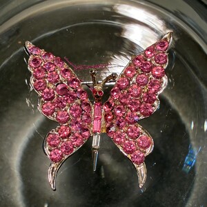 May include: A butterfly-shaped brooch featuring numerous round pink rhinestones covering the wings. The central body is composed of a vertical pink baguette stone and a clear baguette stone at the base. The metal frame is silver-toned.