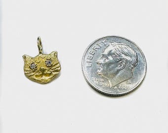 May include: A gold-colored cat pendant with two small, sparkling gemstones for eyes. The pendant is next to a U.S. dime coin for size comparison. The pendant has a textured surface and a loop for a chain.