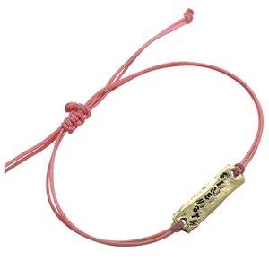 Puede incluir: Una pulsera de cordón rosa con un dije rectangular de color dorado. El dije tiene texto grabado. La pulsera está atada con un nudo para ajustarla.
