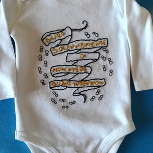 May include: A white long-sleeve baby bodysuit featuring black and gold embroidered text on a ribbon design. The text reads: GIURO SOLENNEMENTE DI NON AVERE BUONE INTENZIONI. Small black embroidered footprints surround the central ribbon design.
