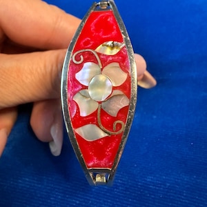 Peut inclure: Un bracelet articulé de couleur argentée doté d'un panneau central ovale allongé rempli d'émail rouge vif. Le centre est orné d'un motif floral composé d'incrustations de nacre blanche irisée, accentué par de fines tiges en métal argenté.