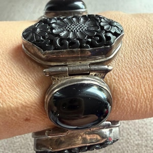 May include: A hinged bracelet featuring alternating links of polished black oval stones and rectangular black carved floral panels set in silver-toned metal. The design showcases intricate petal details on the carved sections and smooth surfaces.