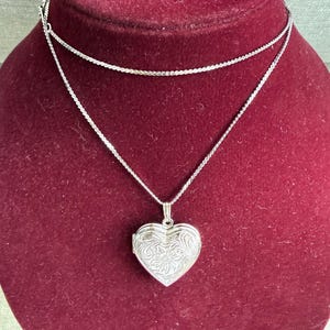 May include: A silver-toned heart-shaped locket pendant suspended from a delicate, thin silver-toned chain. The locket features an intricate, engraved floral pattern across its front surface, showcasing a classic, symmetrical heart design.