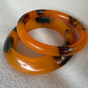 Puede incluir: Dos brazaletes circulares con una base translúcida de color ámbar y patrones moteados en marrón oscuro y negro. Las pulseras tienen un acabado liso y pulido con un perfil redondeado, creando un diseño de aro continuo para la muñeca.