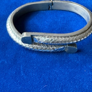 Peut inclure: Un bracelet jonc articulé en métal argenté présentant un motif texturé en forme d'écailles sur toute la surface extérieure. Le design est de style croisé avec deux embouts métalliques arrondis et lisses, ainsi qu'une charnière visible.