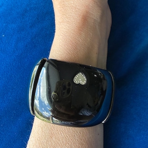 May include: A wide, glossy black cuff bracelet featuring a small, silver-toned heart-shaped embellishment centered on the front surface. The bracelet has a smooth, reflective finish and is constructed with a curved, solid band design.