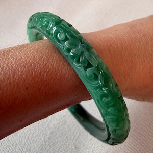 May include: A translucent emerald green bangle bracelet featuring a continuous carved relief pattern of stylised flowers and swirling vine motifs. The circular accessory has a polished, smooth surface with varying shades of deep green.