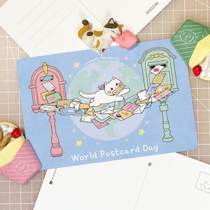 May include: A World Postcard Day postcard featuring a white cat carrying mail between a pink and a green mailbox over a globe. Two cat-themed crepe-shaped memo holders are included, one pink and one green, along with two blank postcards.