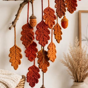 May include: A wall hanging featuring a natural wooden branch with multiple crocheted oak leaves and acorns suspended by yarn. The leaves and acorns are crafted in various shades of orange, red, and brown, creating a layered, cascading arrangement.